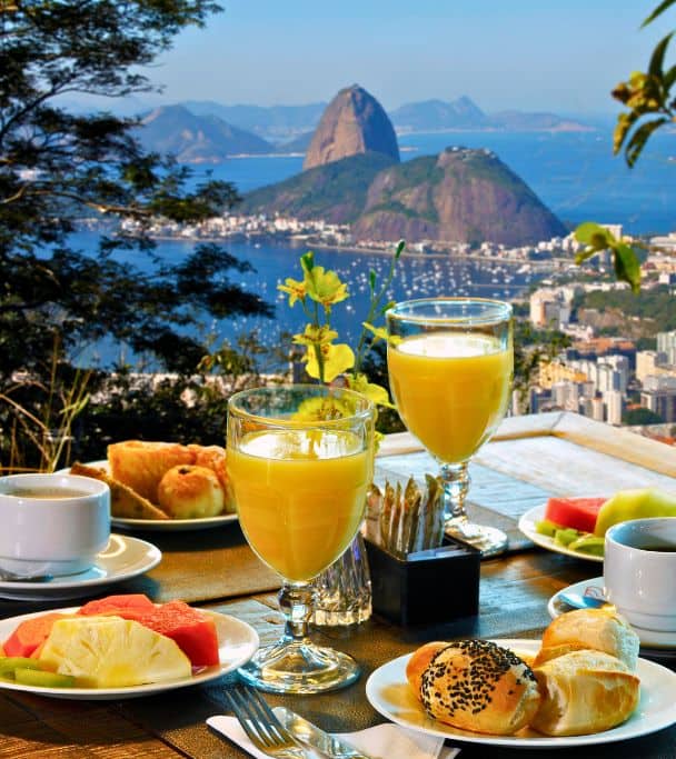 A table of pastries, fruit and orange juice, with a view of Rio de Janeiro in the background. Photo by ribeirorocha via Canva.com.