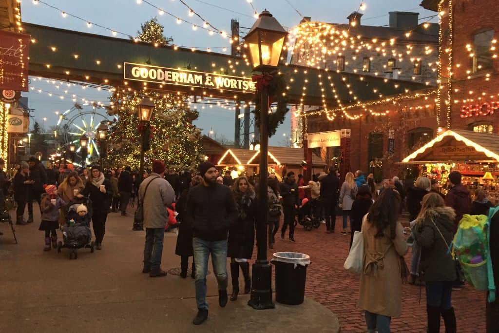 People visiting the Distillery District's winter market, with Christmas lights decorating the square and the famous Gooderham and Worts sign in the background