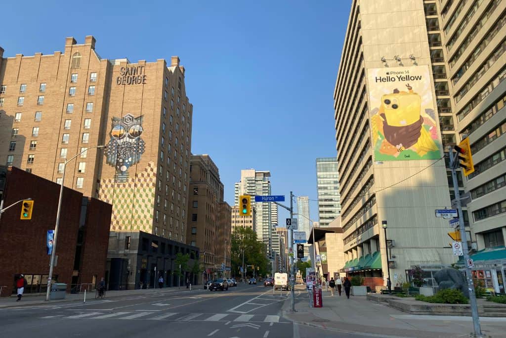 An intersection on Bloor Street, with mid-rise buildings on each side of the road displaying murals and a blue sky in the background