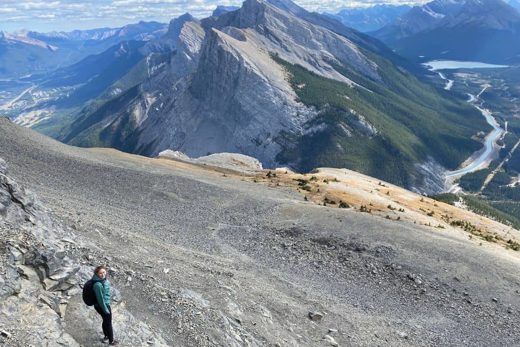 A women who does not regret moving to Canada, standing on the side of a mountain, with the view of a river in the valley below, and more mountains in the background