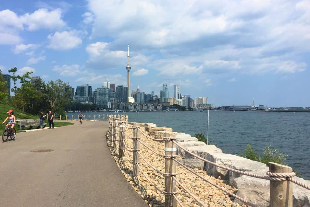 A walkway along Lake Ontario, with some cyclists, and a view of the Toronto Skyline in the background.