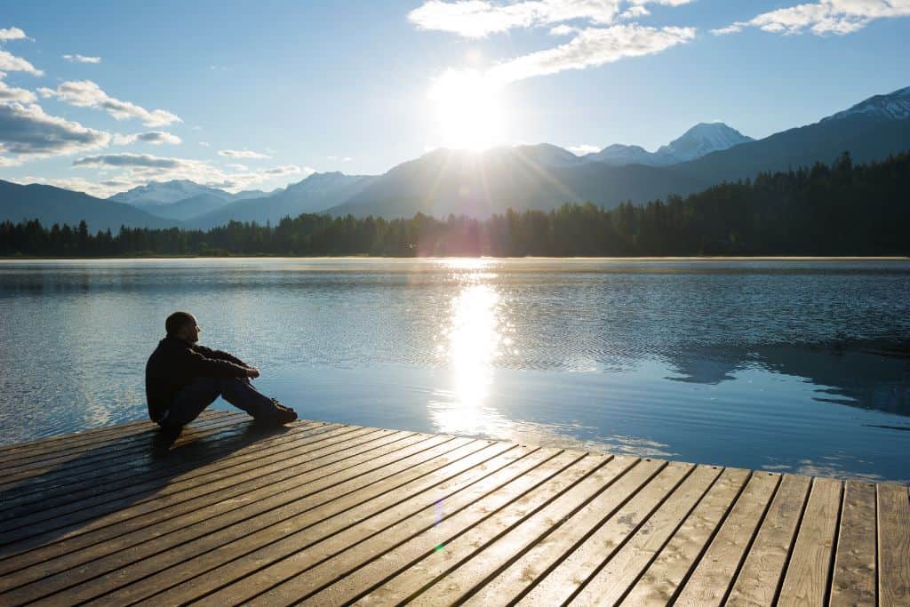 A man sitting alone on a pier at a lake, deep in thought, with mountains in the background and the sun reflecting in the water.