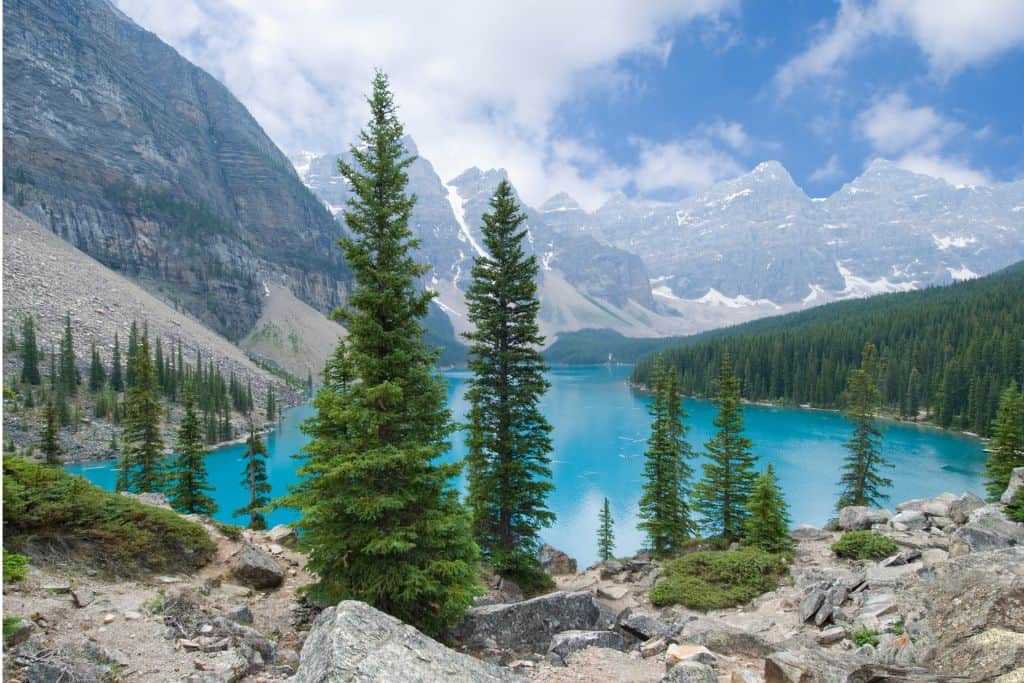 A view of bright blue Moraine Lake through several green trees, with mountains in the backgrounds, partially covered by clouds. This picturesque setting is a reason you won't regret moving to Canada.