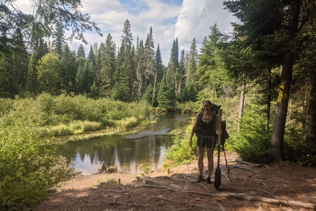 A women who doesn't regret moving to Canada, carrying a hiking pack and two paddles standing in-front of a small lake, surrounded by green forest