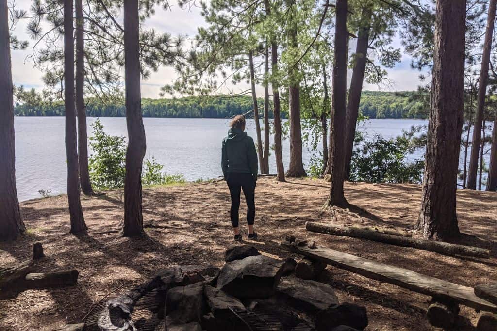 A woman wearing a green jacket, standing among some trees in a lakeside campsite, with the blue water visible in the background