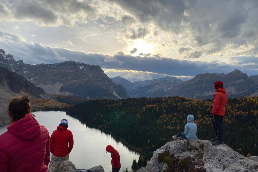 A group of hikers sitting on some rocks overlooking a lake with mountains in the background on a cloudy day.