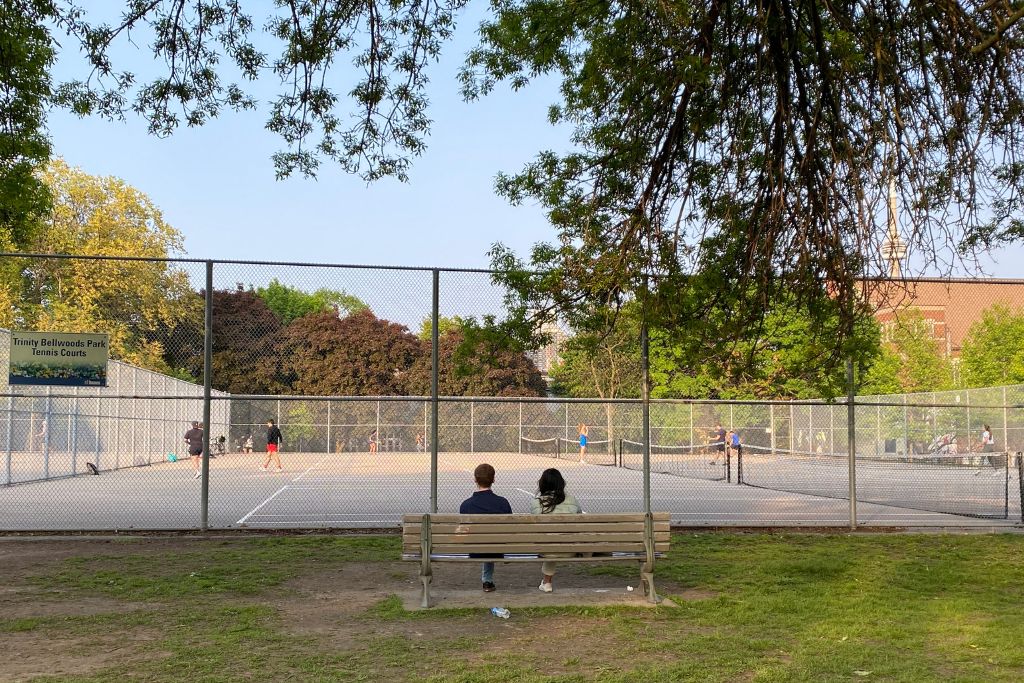 A couple sitting on a park bench in front of an enclosed area with four tennis courts, watching the tennis players.