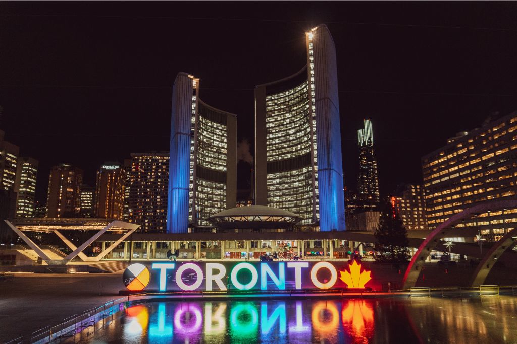The Toronto sign lit up in with multiple coloured lights reflecting in a pond of water at night, in a relatively safe area in Toronto.