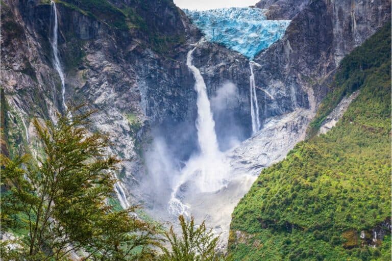 A blue glacier hanging over the edge of a cliff with several small waterfalls cascading down in Queulat National Park