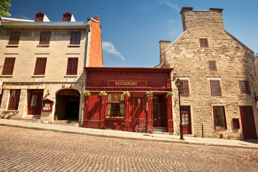 A historic red restaurant sandwiched between two grey stone buildings on a cobble stone street in old Montreal