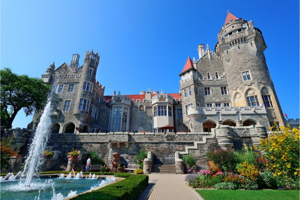The front of the gothic revival castle 'Casa Loma' in Toronto's midtown, with a blue fountain to the left and a colourful garden to the right.