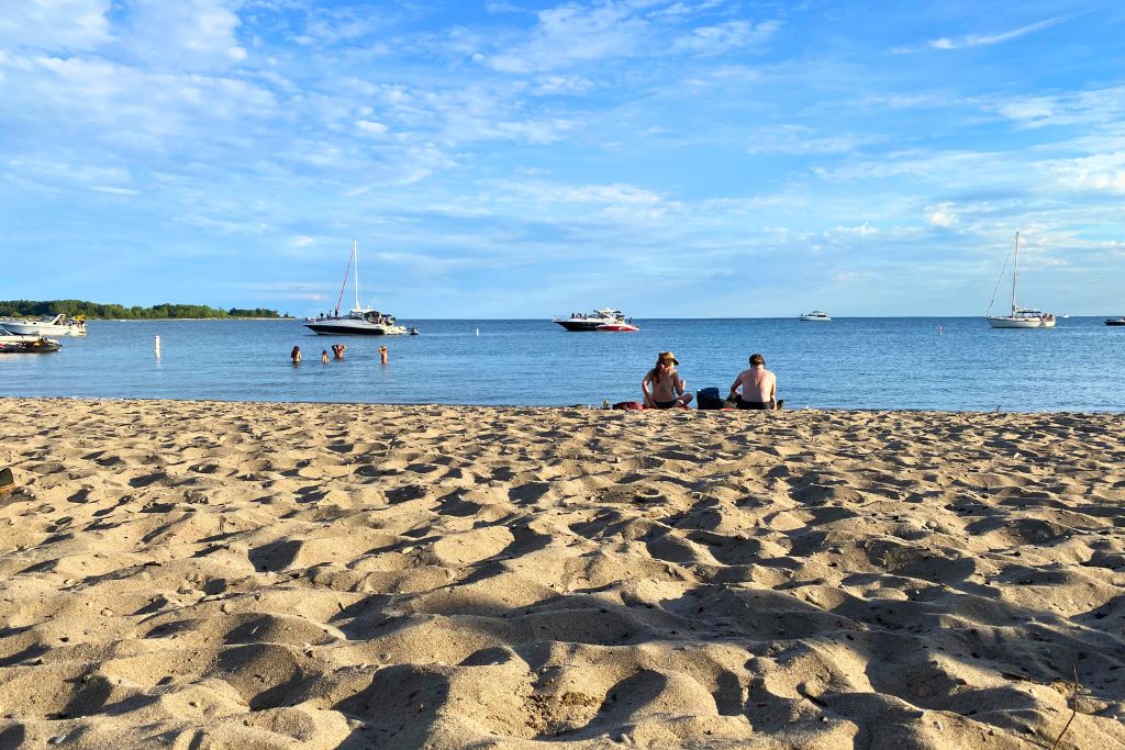 A couple sitting on a beach at the Toronto Islands with boats on the water in the background.